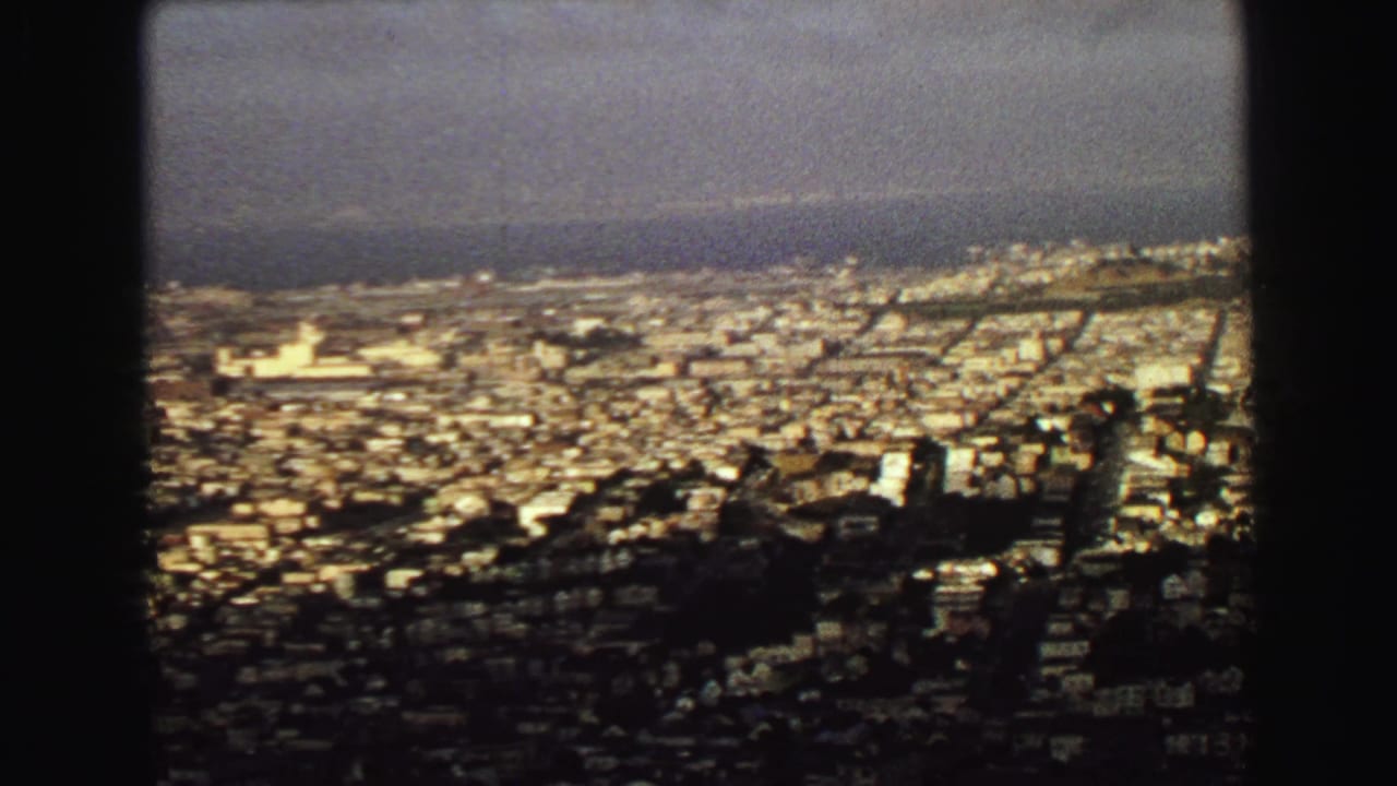 High view over dense city buildings toward distant water, framed by dark window edges.