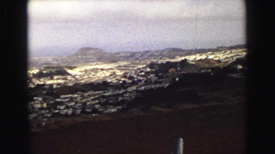 1968 8mm hilltop view in Vancouver, with a brown foreground field, cemetery rows, low buildings, distant hills, and hazy sky.