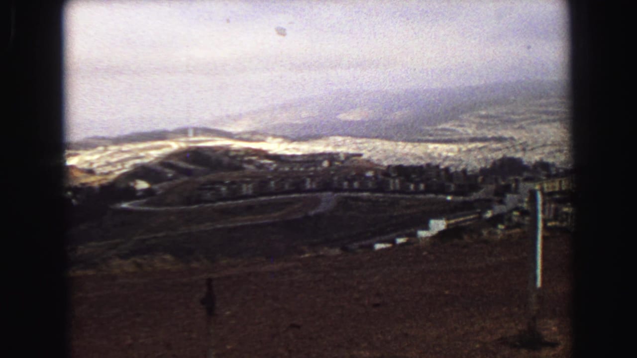 Elevated 1968 Vancouver view with cemetery markers, a curved road or path, hillside buildings, brown ground, and distant city haze.