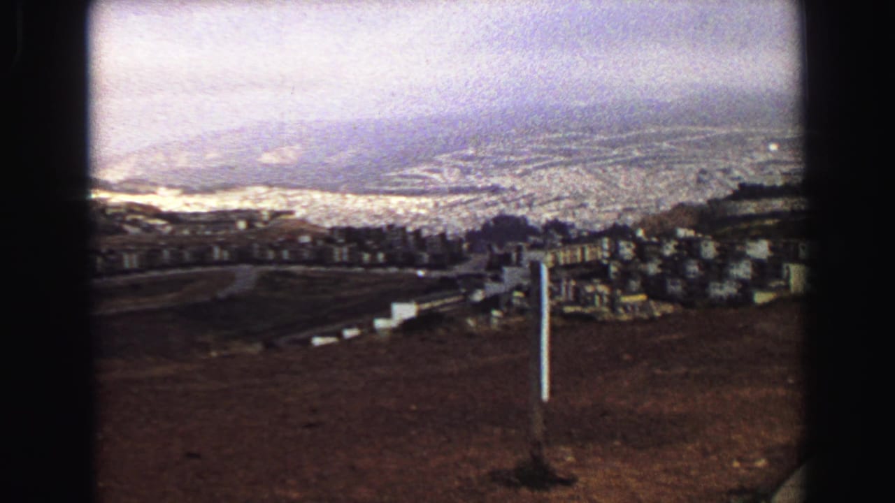 Late pan frame from Vancouver in 1968, with brown hillside foreground, cemetery markers, clustered buildings, distant urban grid, and hazy horizon.
