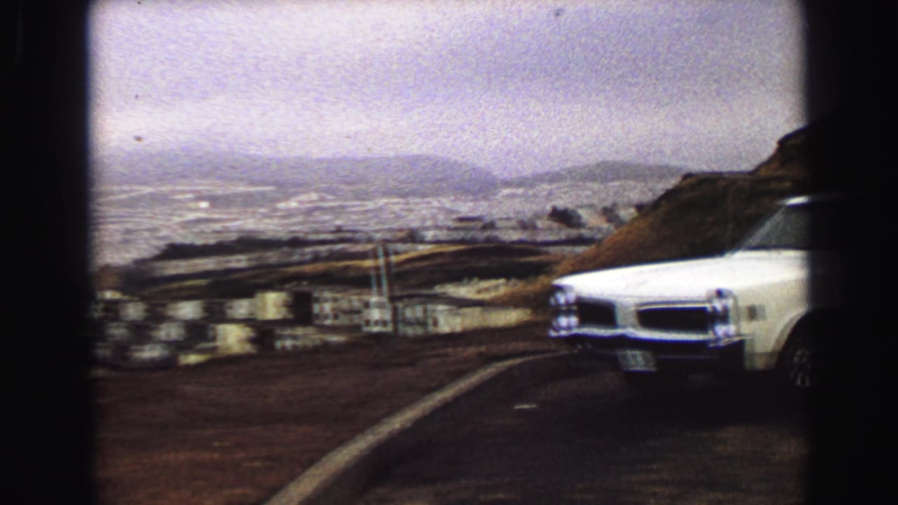 A white late-1960s style car appears beside the hillside road, with Vancouver and the coastline in the distance.