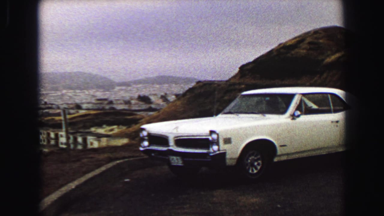 White classic coupe parked at a Vancouver hillside overlook in 1968, with the city, water, and low hills behind it.