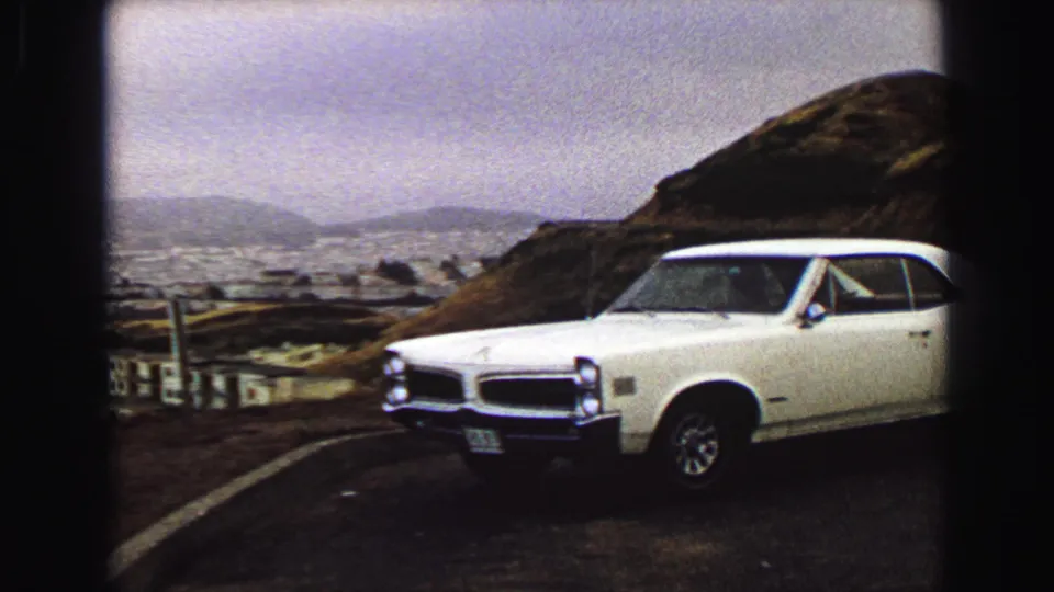 White classic coupe parked at a Vancouver hillside overlook in 1968, with the city, water, and low hills behind it.