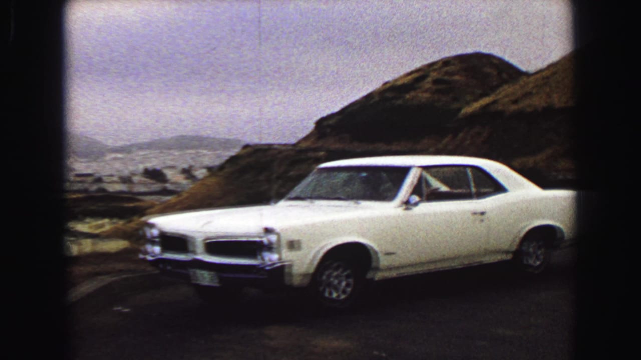 White classic car parked beside a rocky coastal hillside under a hazy sky.