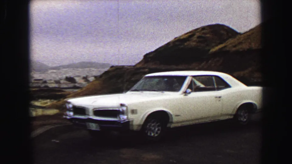 1968 8mm frame of a white classic car parked on an elevated roadside near Vancouver, with rocky hillside and hazy landscape behind it.