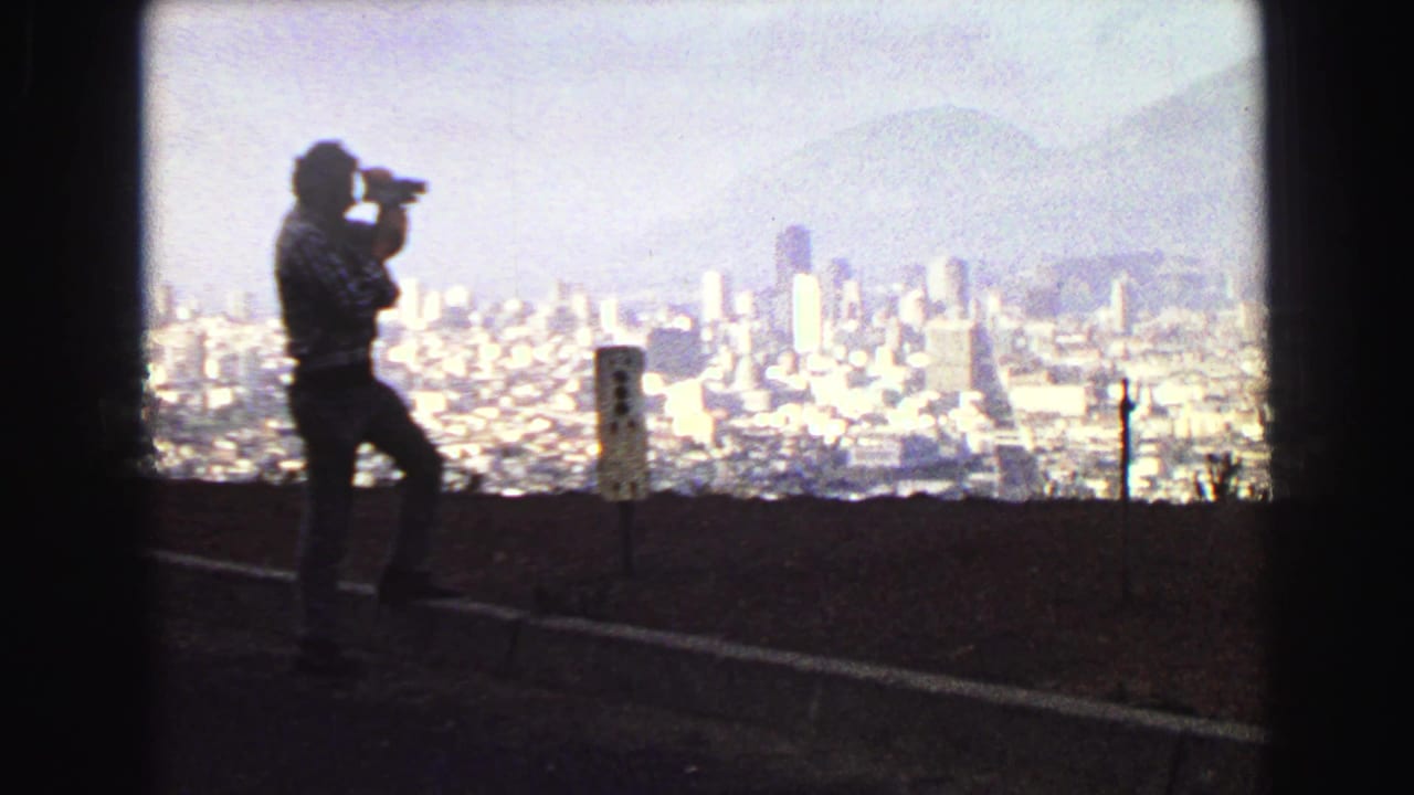 A man in silhouette photographs the Vancouver skyline from an elevated roadside viewpoint in this 1968 8mm home-movie frame.