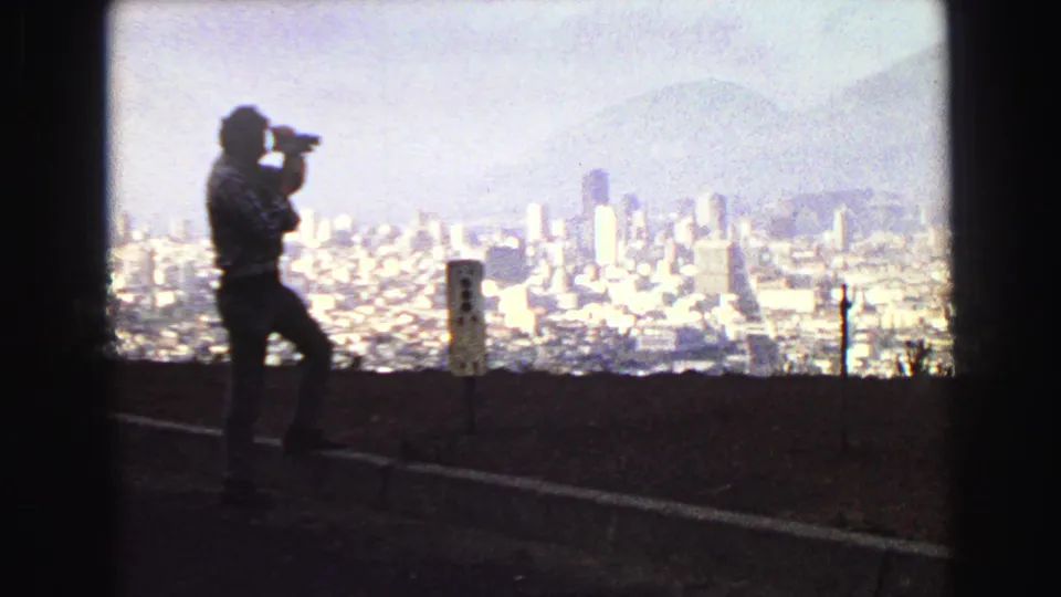 A man in silhouette photographs the Vancouver skyline from an elevated roadside viewpoint in this 1968 8mm home-movie frame.