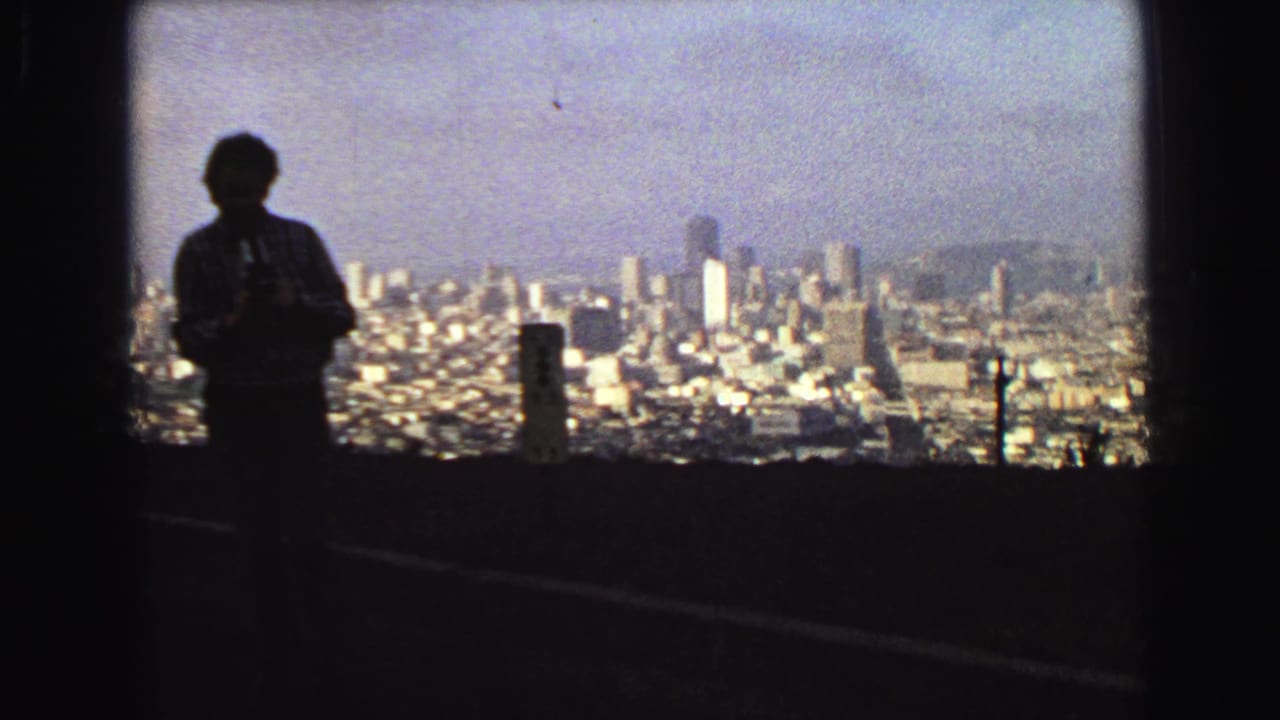 The silhouetted traveler stands near roadside posts with Vancouver's dense urban view spread below in grainy 1968 home-movie footage.