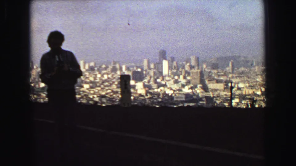 The silhouetted traveler stands near roadside posts with Vancouver's dense urban view spread below in grainy 1968 home-movie footage.
