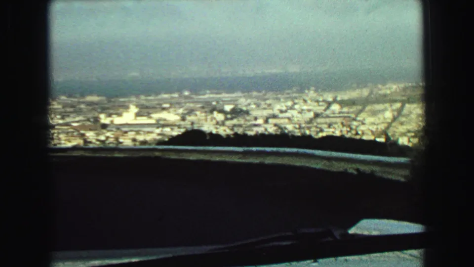 1968 Vancouver cityscape seen from inside a vehicle, with a windshield wiper, railing, hillside edge, haze, and distant water.