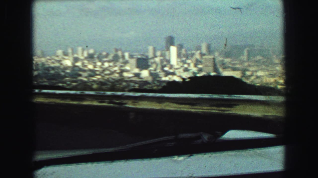 Elevated 1968 Vancouver skyline view with taller downtown buildings, hillside foreground, haze, railing, dashboard, and windshield wiper.