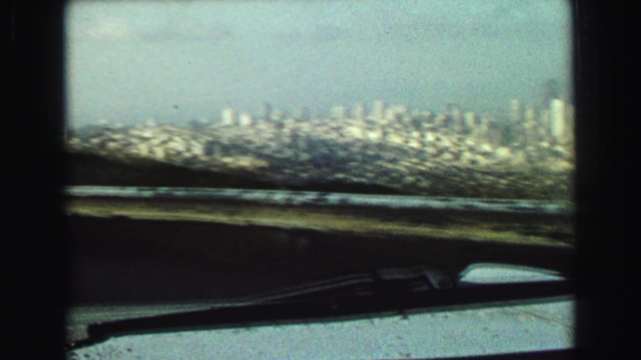 1968 Vancouver-area windshield view with a distant hazy skyline, dark dashboard, and windshield wiper visible from inside a moving vehicle.