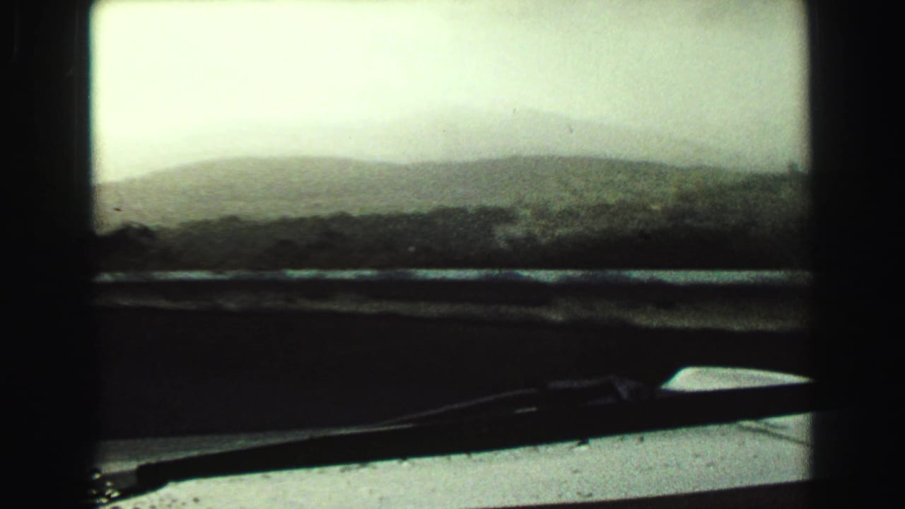 1968 Vancouver-area view from inside a moving vehicle, showing a soft mountain silhouette, muted sky, windshield frame, dashboard, and wiper.