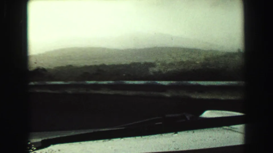 1968 Vancouver-area view from inside a moving vehicle, showing a soft mountain silhouette, muted sky, windshield frame, dashboard, and wiper.