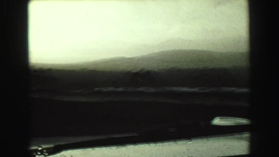 1968 8mm frame of a hazy roadside or landscape view from inside a moving car, with windshield edge and soft exposure.