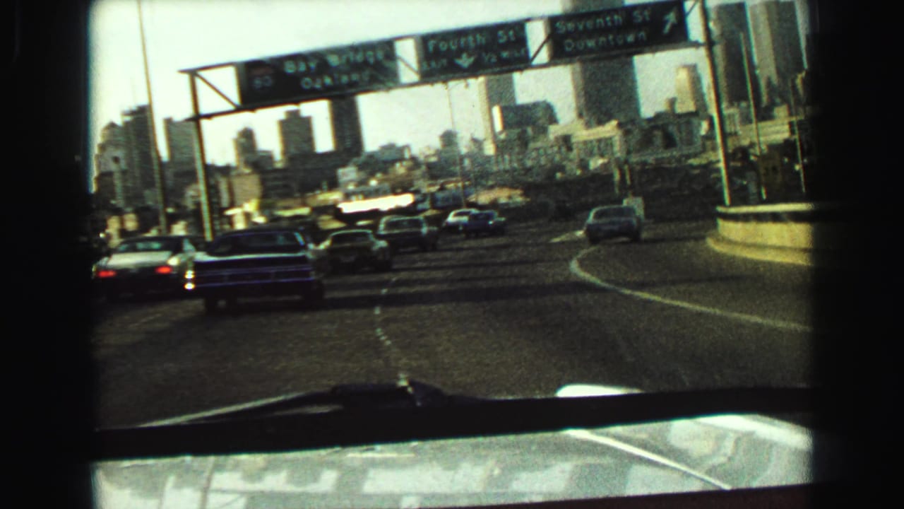 Highway traffic approaches overhead downtown road signs and a distant skyline.