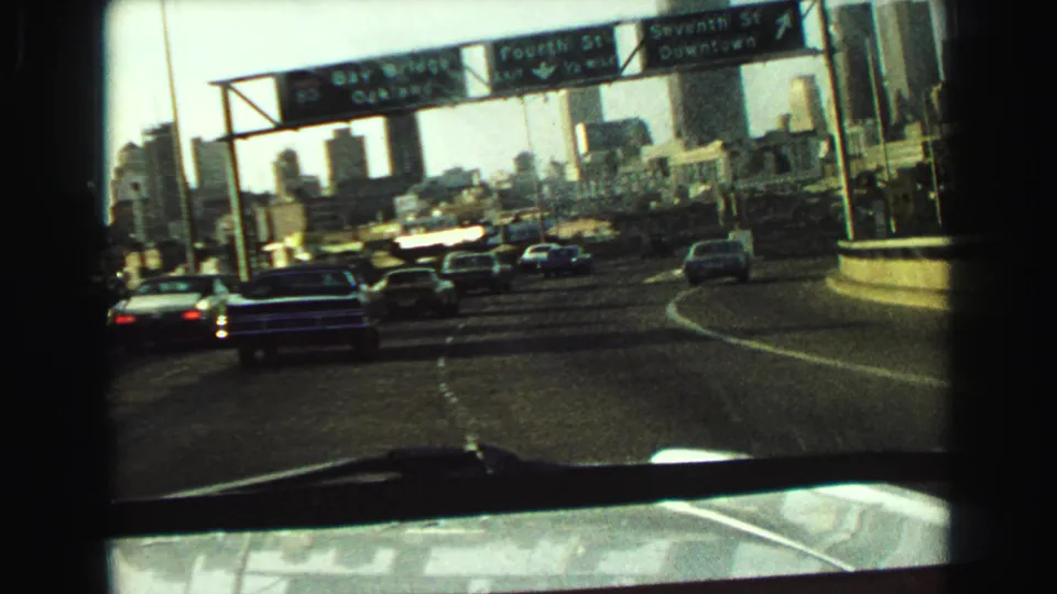 1968 8mm view from inside a car of multi-lane highway traffic, overhead signs, and high-rises in the distance.