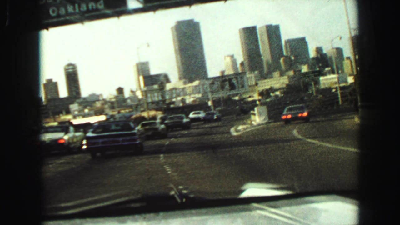1968 8mm passenger-view frame of classic cars on a divided highway with mid-century high-rises ahead.