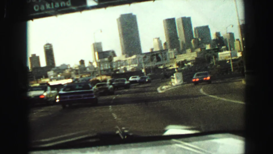 1968 8mm passenger-view frame of classic cars on a divided highway with mid-century high-rises ahead.