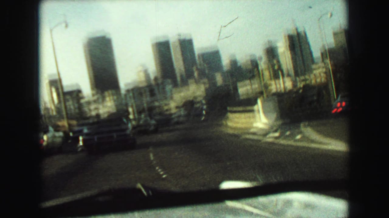 1968 8mm frame from a moving car showing blurred traffic lanes, roadside lights, and downtown towers through the windshield.