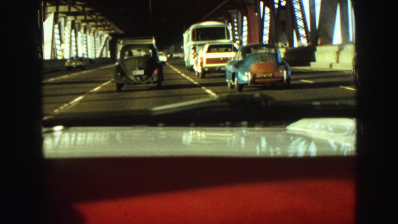 1968 Vancouver bridge traffic filmed from inside a moving car, with classic cars ahead and steel trusswork casting shadows.