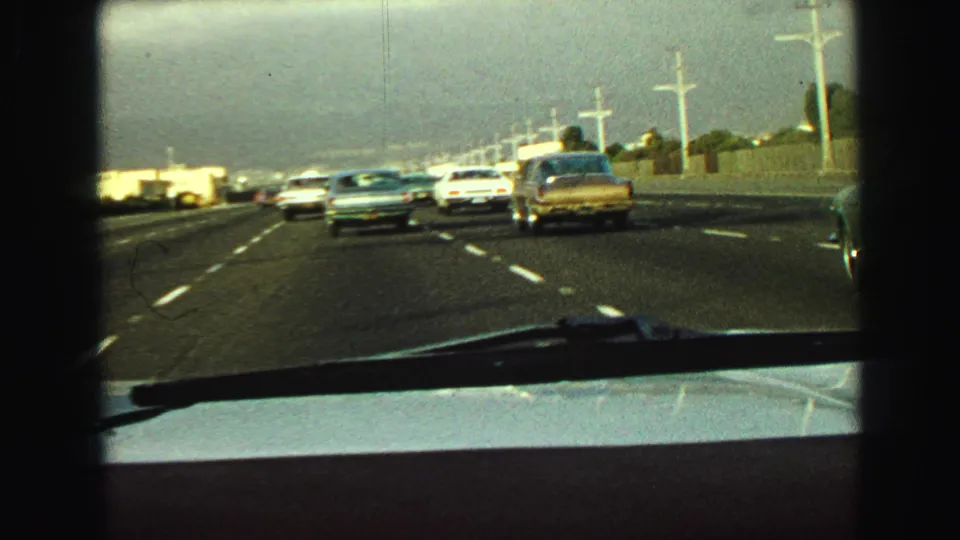 In-car 8mm view of late-1960s freeway traffic near Vancouver, with a white car, blue sedan, and gold sedan visible ahead.