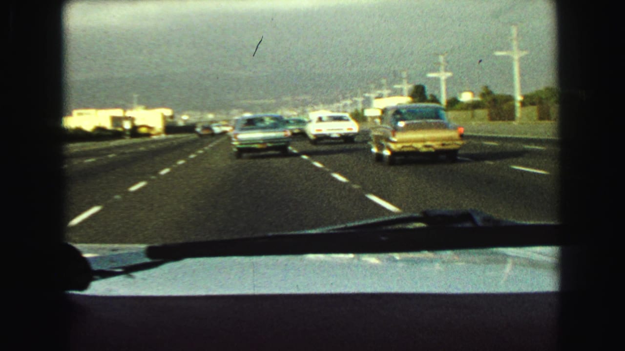 Windshield-shot 1968 freeway scene with a gold sedan at right, other period cars ahead, and visible road texture and lane stripes.