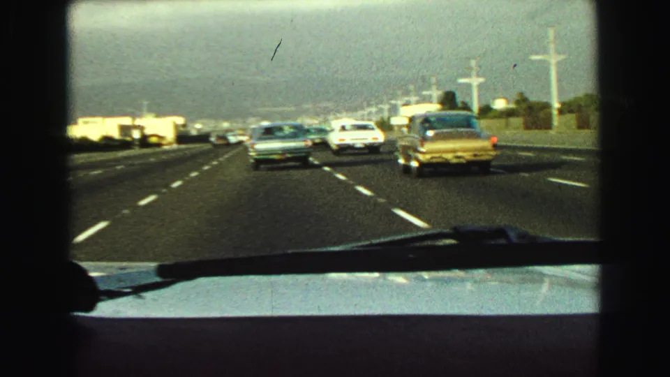 Windshield-shot 1968 freeway scene with a gold sedan at right, other period cars ahead, and visible road texture and lane stripes.