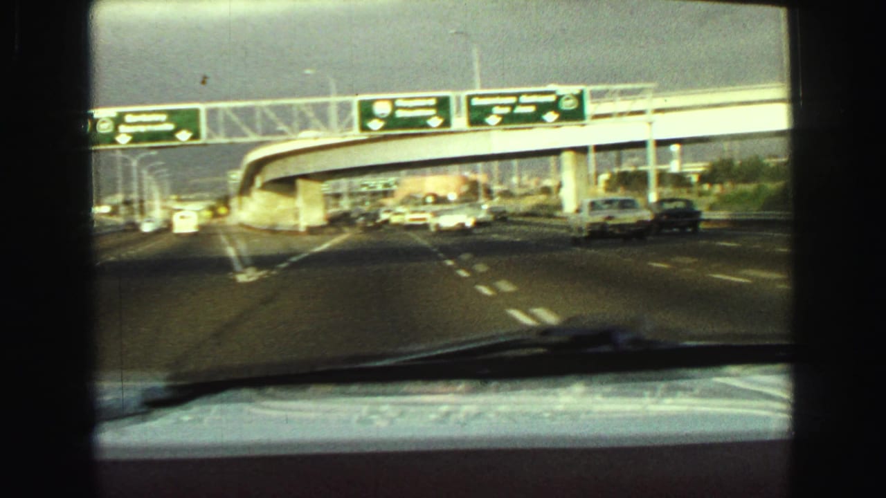 1968 Vancouver highway approach filmed through a windshield, showing cars, an overpass, and overhead directional signs.