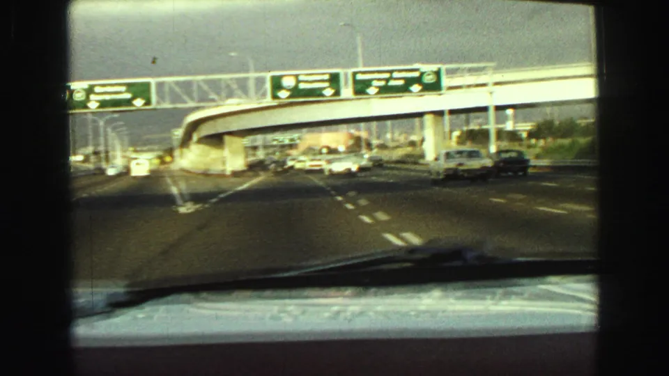 1968 Vancouver highway approach filmed through a windshield, showing cars, an overpass, and overhead directional signs.