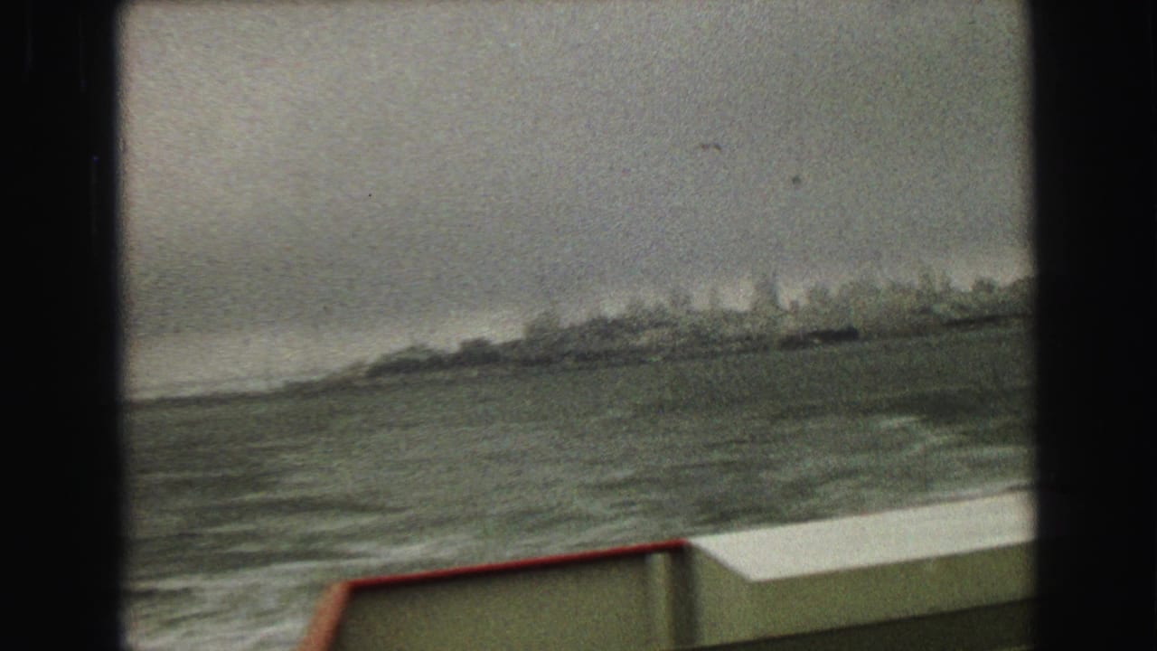 Hazy 8mm view from a boat showing rough water, a low distant shore or headland, and the red rail along the deck.