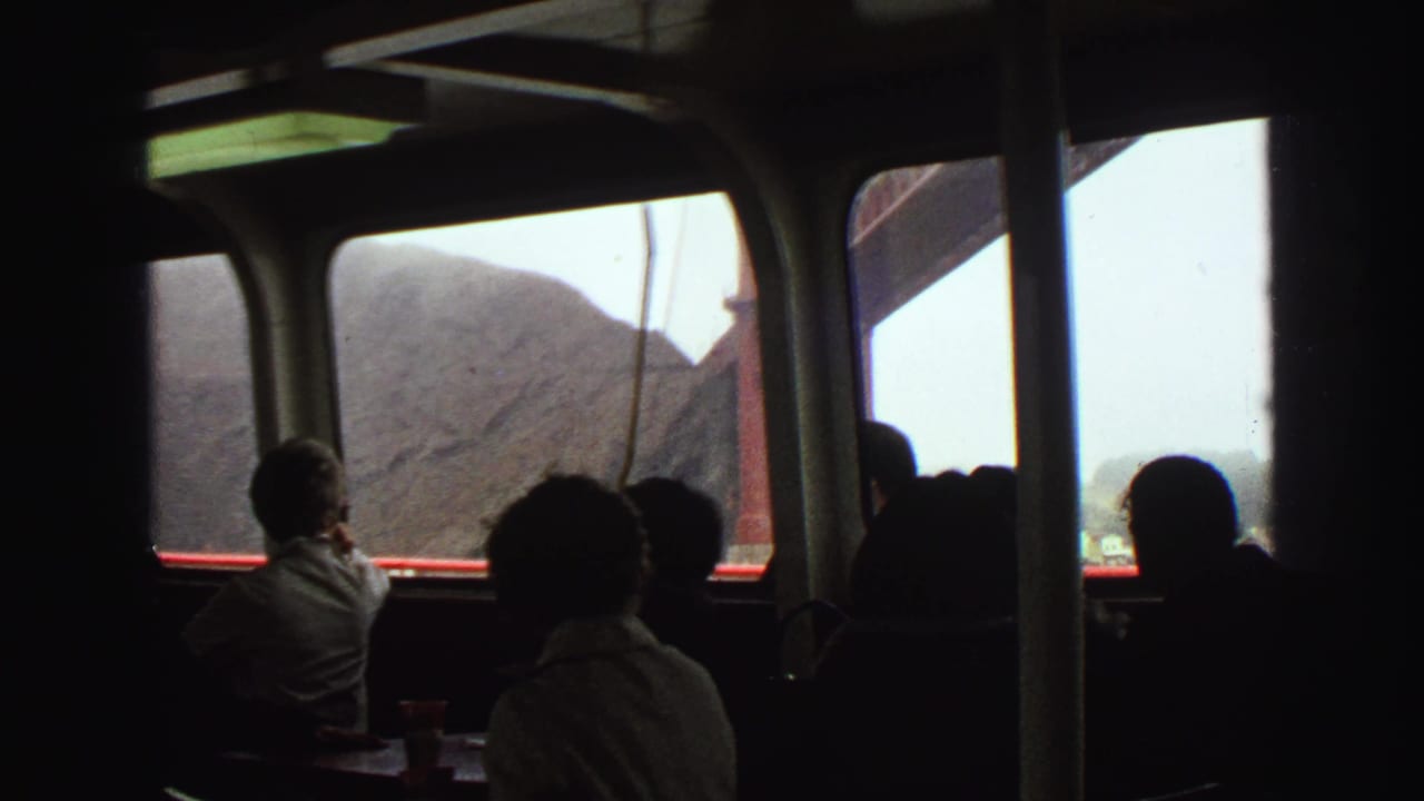 1968 8mm travel frame with silhouetted passengers inside a boat cabin, looking toward hills and a red bridge structure through the windows.