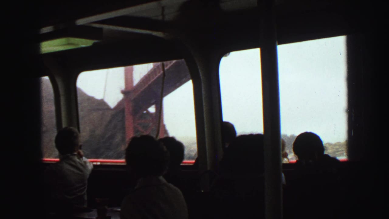 1968 8mm frame showing several passengers in a dark boat interior beside windows, with a red bridge support and hillside outside.