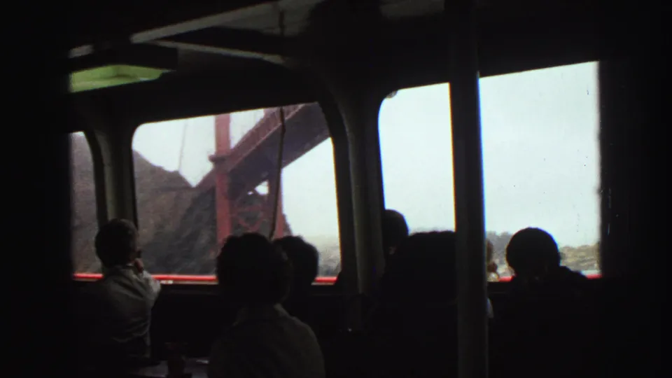 1968 8mm frame showing several passengers in a dark boat interior beside windows, with a red bridge support and hillside outside.