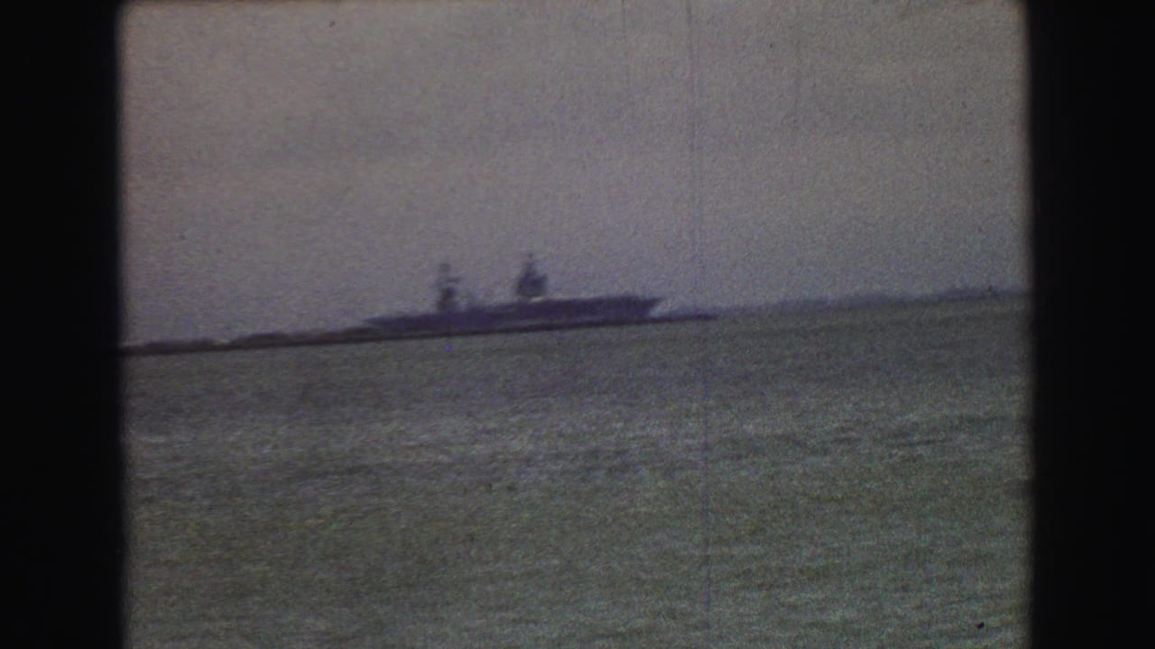 1968 8mm frame showing a distant ship on open water, filmed with heavy grain, soft focus, and muted color.