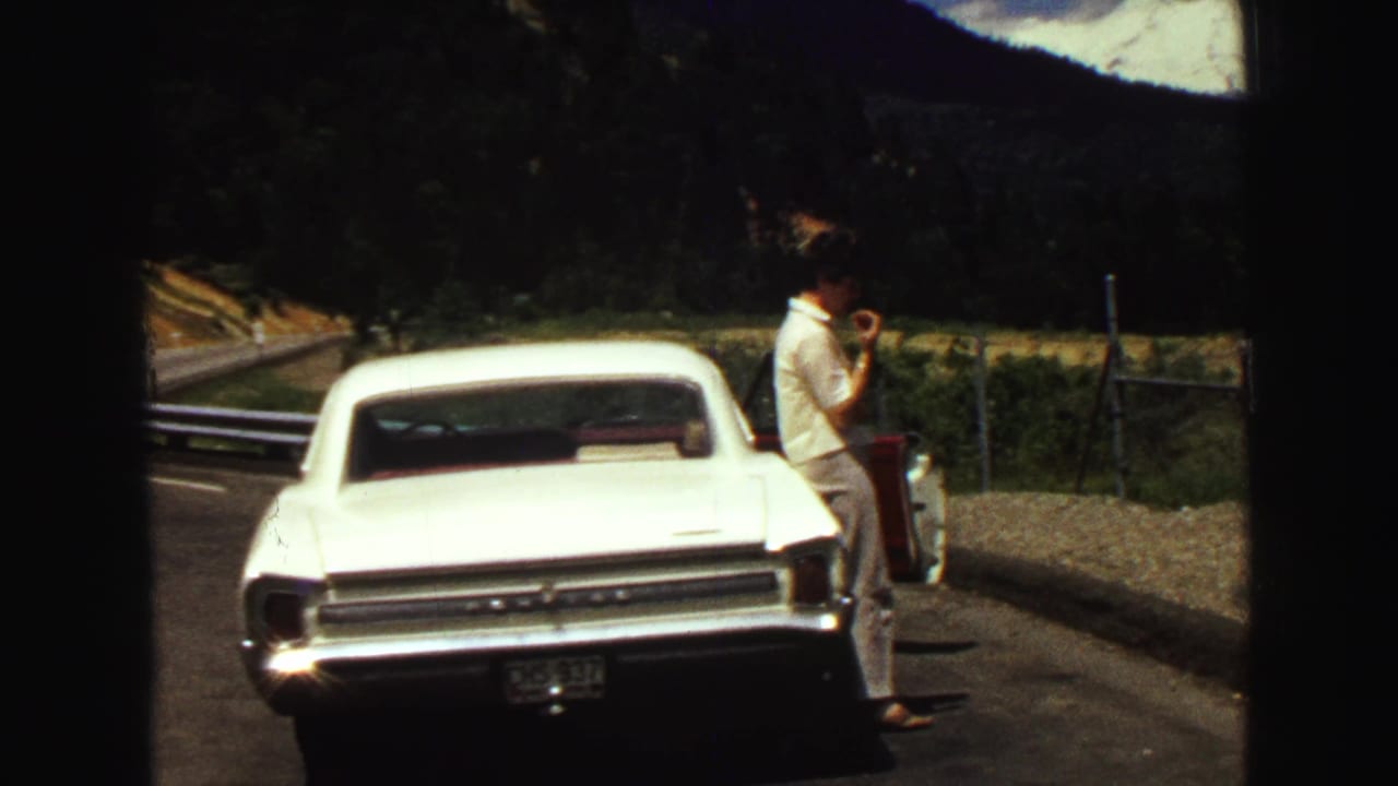 1968 Vancouver-area roadside scene with a man beside a white classic car, fencing, forested slopes, and mountains in the background.