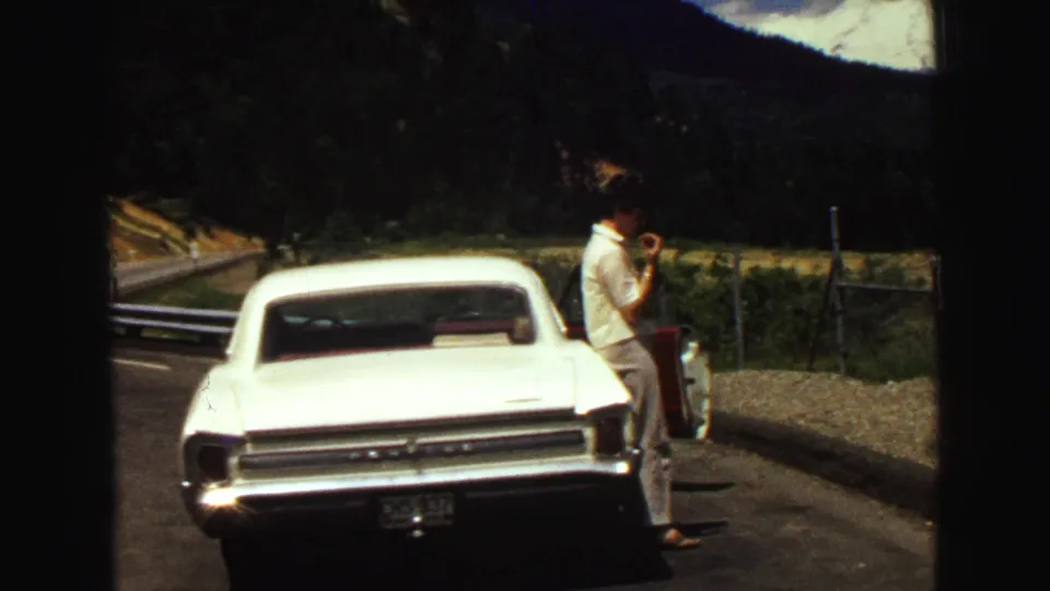 1968 Vancouver-area roadside scene with a man beside a white classic car, fencing, forested slopes, and mountains in the background.