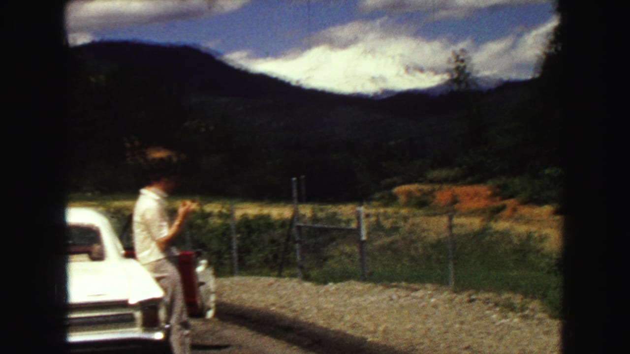 A man stands near a white classic car on a Vancouver-area mountain road, framed by fencing, hills, and dramatic cloud cover.