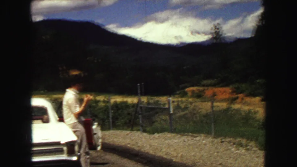 A man stands near a white classic car on a Vancouver-area mountain road, framed by fencing, hills, and dramatic cloud cover.