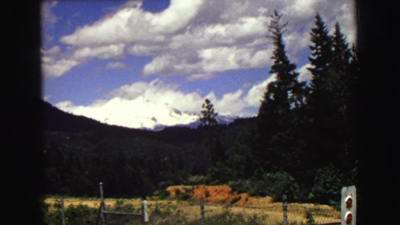 1968 8mm frame of a Vancouver-area snow-capped mountain above forest and roadside fencing under a bright clouded sky.
