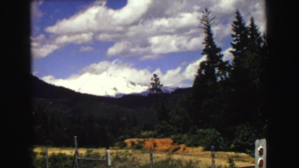 1968 8mm frame of a Vancouver-area snow-capped mountain above forest and roadside fencing under a bright clouded sky.