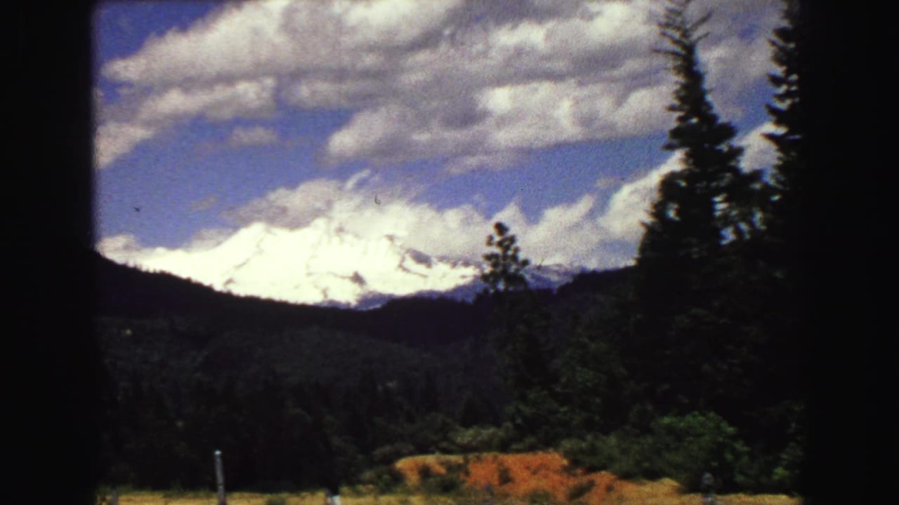 1968 8mm Vancouver-area landscape showing a closer snow-covered mountain view across forest, grass, fence posts, and clouded blue sky.