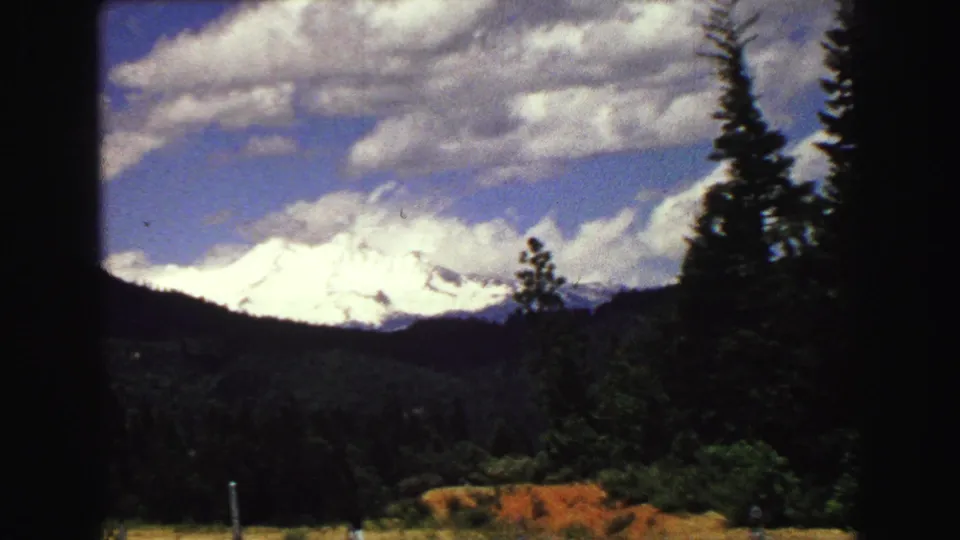 1968 8mm Vancouver-area landscape showing a closer snow-covered mountain view across forest, grass, fence posts, and clouded blue sky.