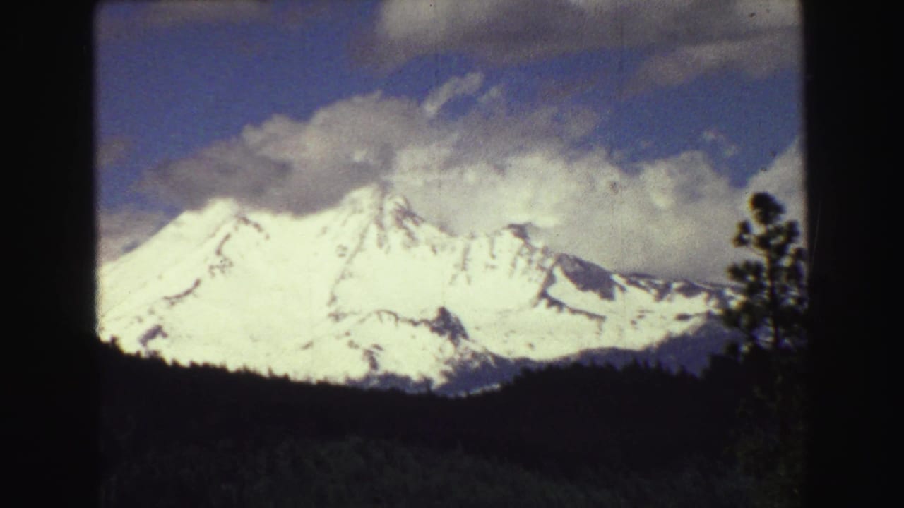 Snow-capped mountain range behind a dark evergreen tree line and black film border.