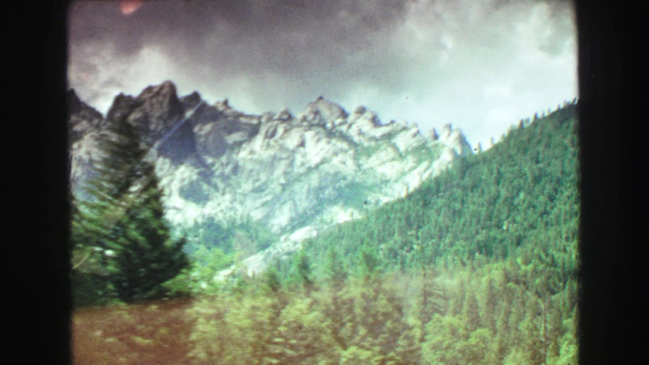 1968 8mm home-movie frame of rocky British Columbia mountain ridges above evergreen forest and cloudy sky.