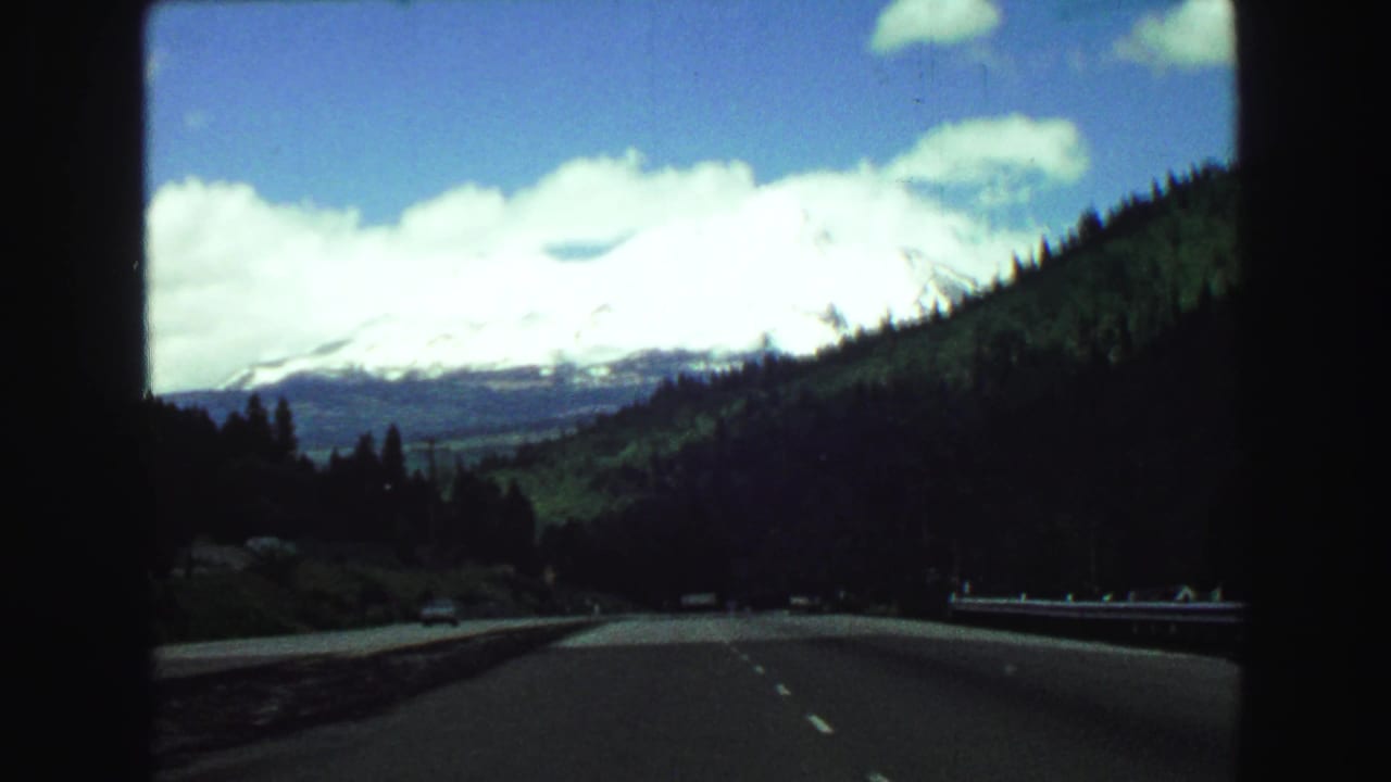 Highway curving through forested mountains toward a bright snow-capped peak.