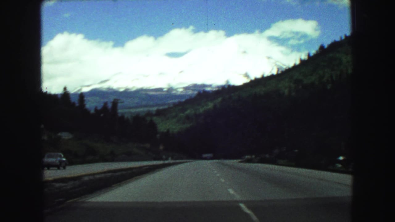 From a moving car, the 8mm frame shows a highway running between forested hills toward a bright mountain range and cloud-filled sky.