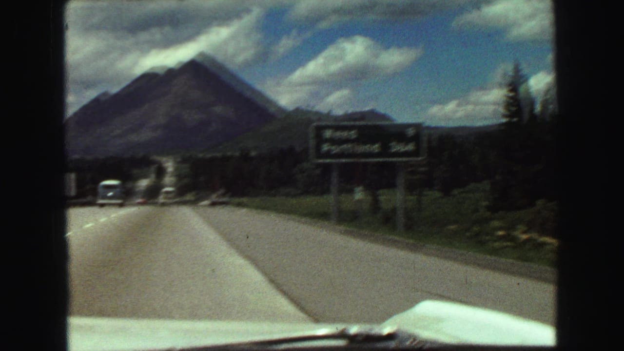 A roadside sign and passing cars appear along a mountain highway, with a dark peak and forested roadside visible in the background.