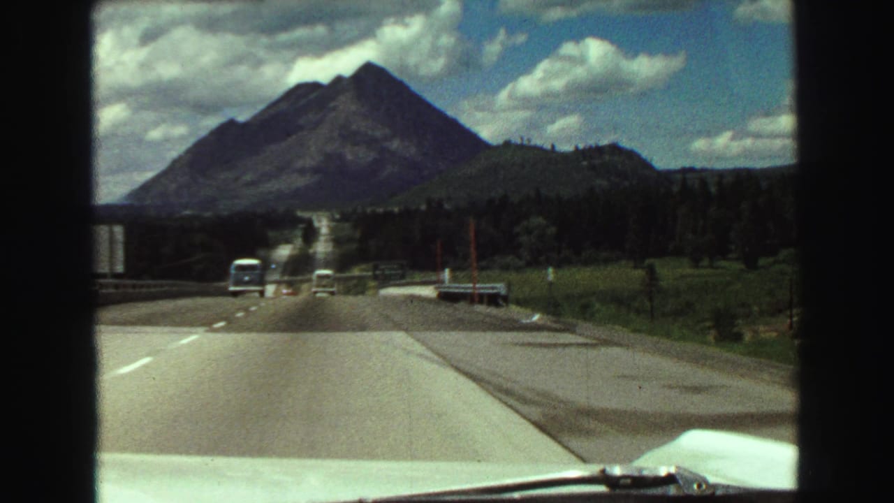 1968 8mm highway view from a moving car, with mountain scenery, traffic, grassland, and the car hood visible.