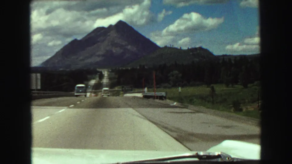 1968 8mm highway view from a moving car, with mountain scenery, traffic, grassland, and the car hood visible.